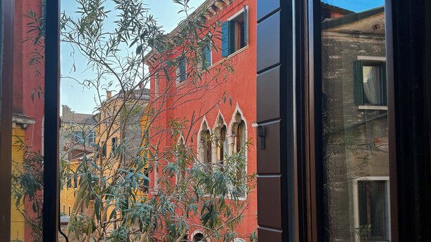 A view through a window of a building with a red facade, a balcony, and some greenery. - SAN VIO PALACE LUXURY APARTMENTS (Venice)