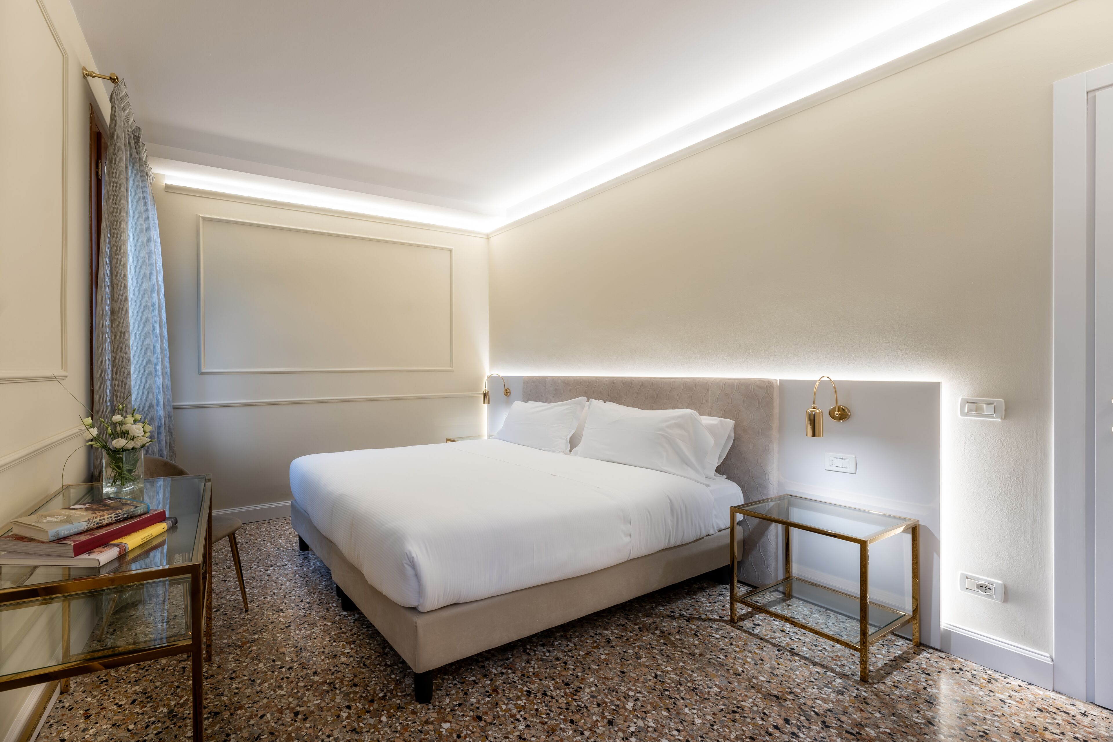 A neatly arranged hotel room with a bed, a glass table with books, a vase of flowers, and a small side table.