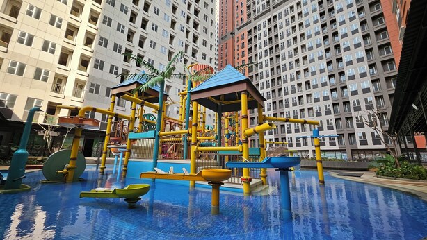 A water play area with slides and climbing structures, surrounded by high-rise residential buildings. - Transpark Juanda Bekasi by 21 Room (Bekasi)