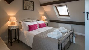 A neatly made bed with white and pink pillows, a quilted bedspread, and a set of towels on the side. There are two bedside tables with lamps, a framed picture on the wall, and a skylight above the bed.