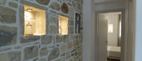 A hallway with a stone wall, a lit shelf with decorative items, and an open door leading to a bedroom.