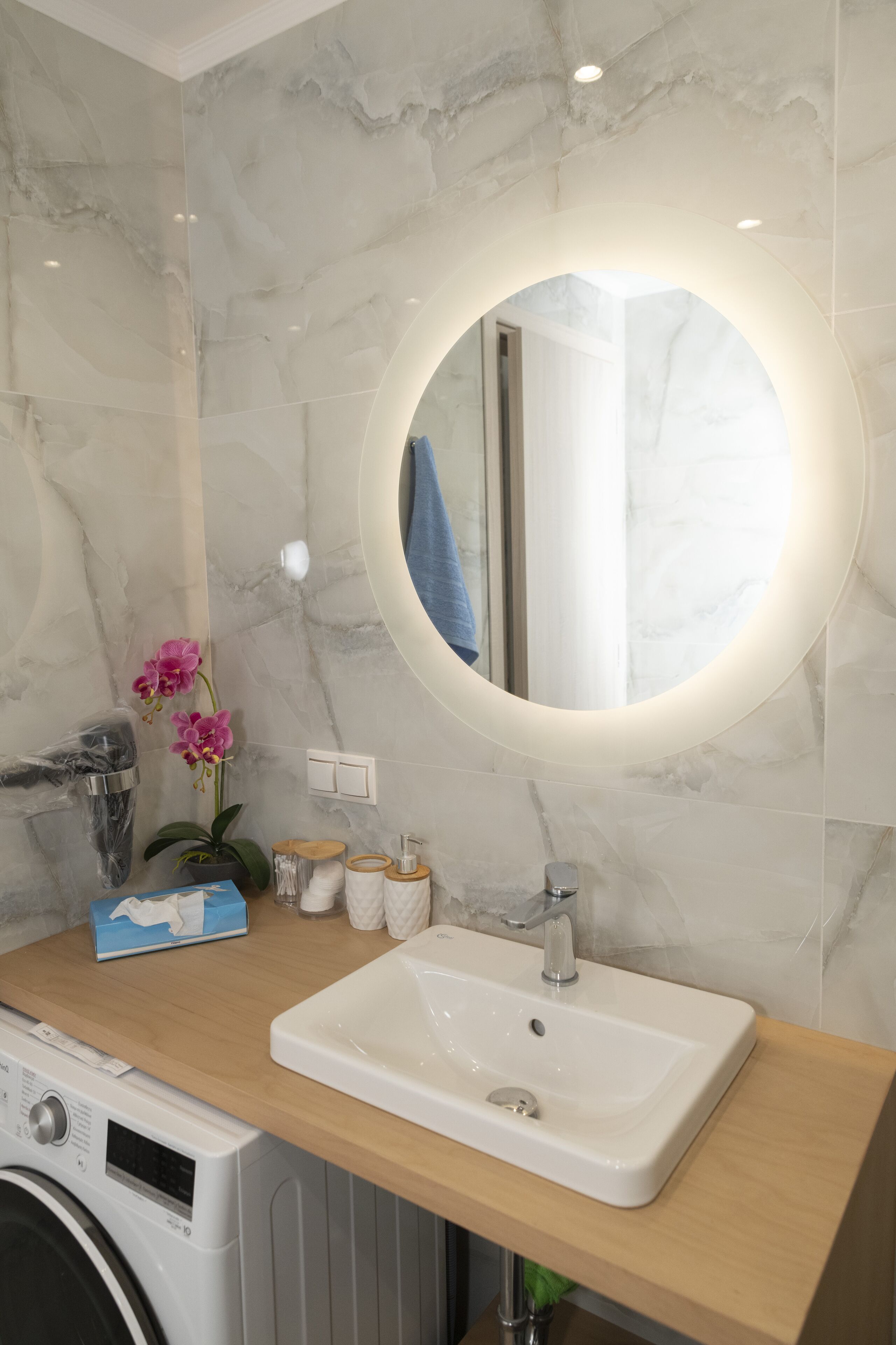 Une salle de bain avec un mur en marbre, un miroir rond, un lavabo, un lave-linge et une orchidée en pot.