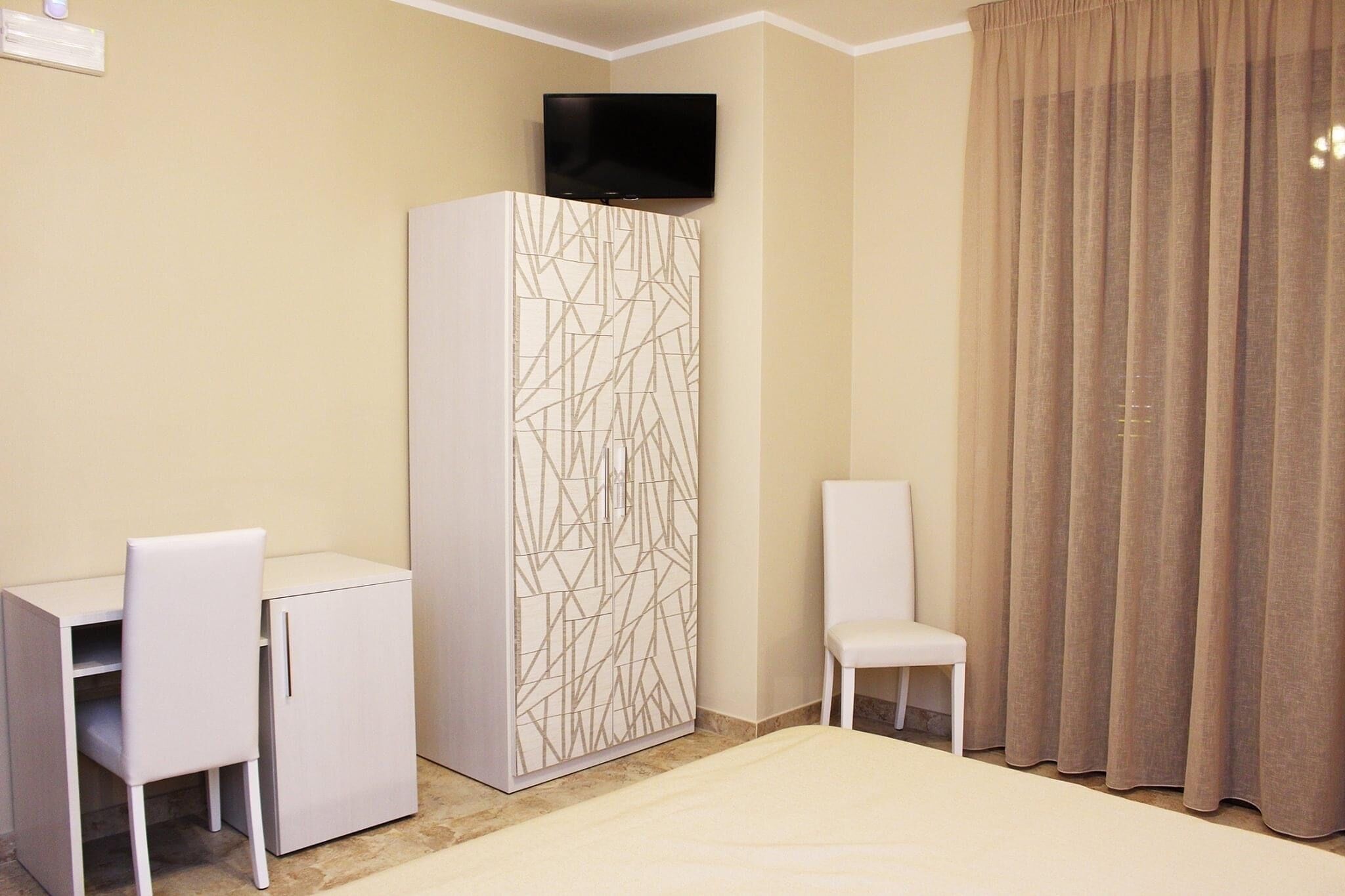 Une chambre d’hôtel comprenant un lit, un bureau avec une chaise, une armoire, un téléviseur fixé au mur et une fenêtre avec des rideaux.