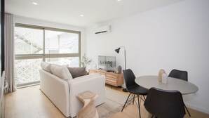 Una sala de estar moderna con un sofá, una mesa de comedor con sillas, un televisor y una escalera visible a través de la ventana.