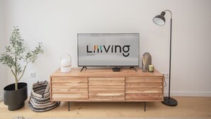 Una sala moderna con un televisor de pantalla plana sobre un mueble de madera, una lámpara de pie, una planta en maceta y un bolso a rayas.