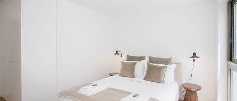 Una cama bien hecha con cubrecama blanco y beige, dos lámparas de noche y una mesita de madera.