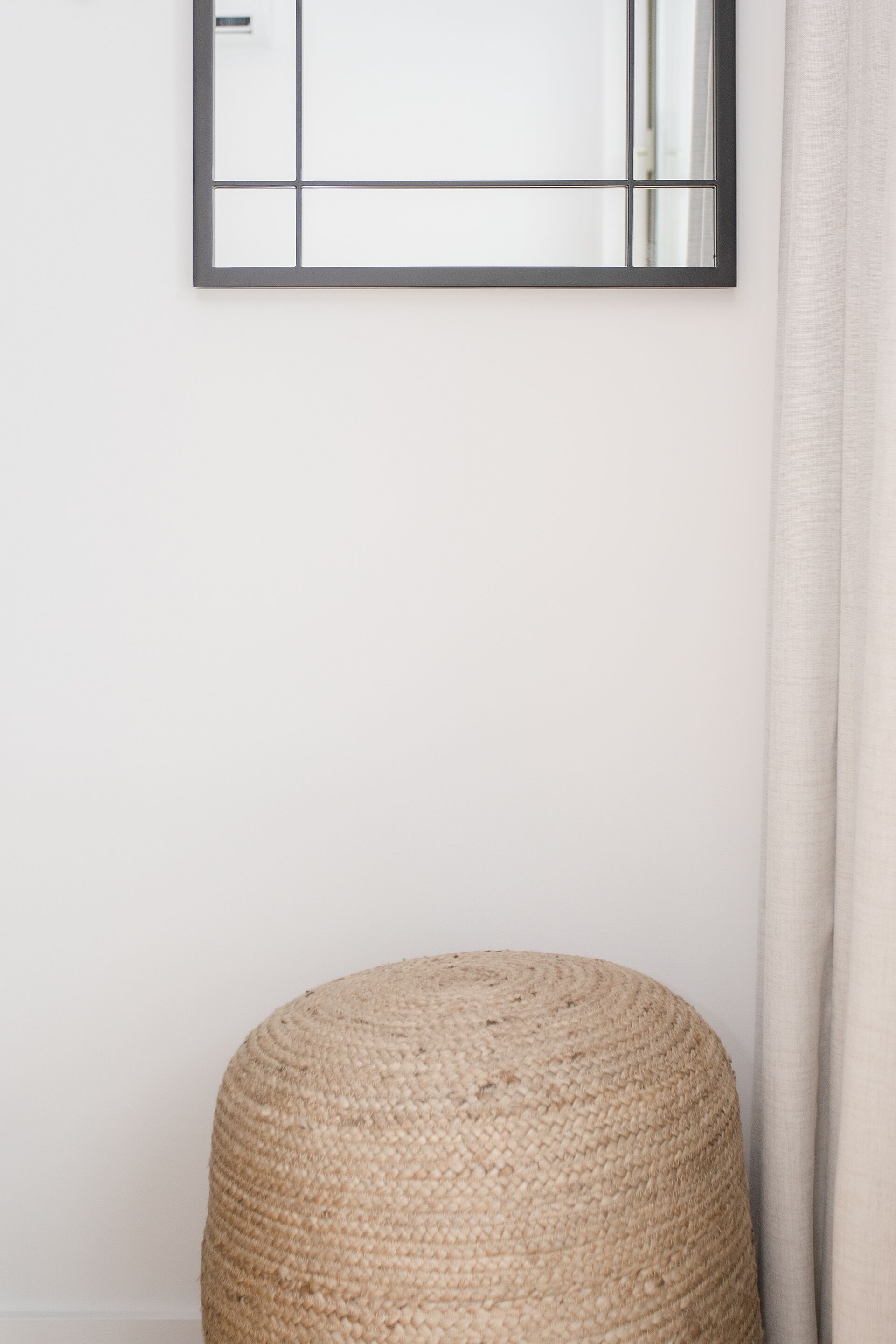 A room with a plain white wall, a framed mirror, and a round woven ottoman.