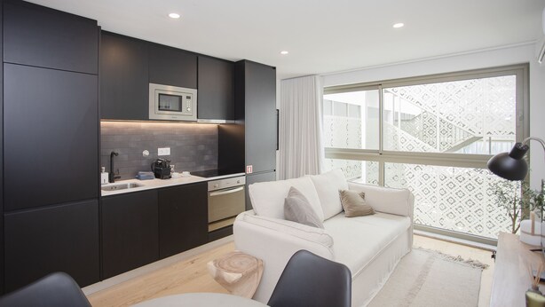 Cocina moderna con armarios oscuros, microondas y pileta. Hay un sofá blanco con almohadones y un ventanal con vista al exterior.