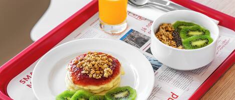 Un plateau de petit-déjeuner avec une assiette de crêpes garnies de fruits et de granola, un verre de jus d’orange et un bol de céréales avec des tranches de kiwi.