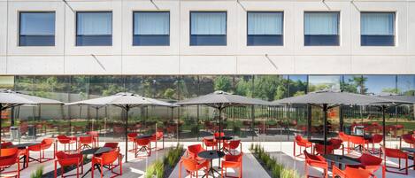 Un área de estar exterior moderna con sillas y mesas rojas, protegida por grandes sombrillas, y un muro de cristal que refleja la vegetación circundante.