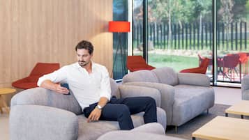 Um homem sentado em um sofá cinza numa sala moderna com parede de madeira e uma janela grande.