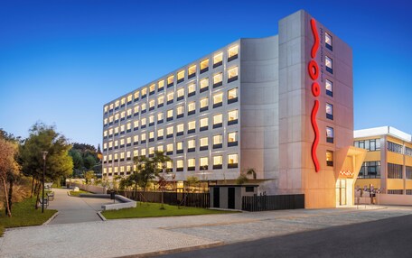 A modern hotel building with a prominent red logo, multiple floors, and a well-maintained outdoor area with pathways and greenery.