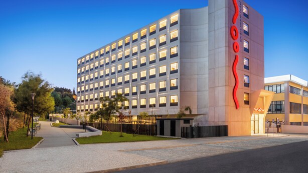 Um hotel moderno com um logotipo vermelho marcante, vários andares e uma área externa bem cuidada, com caminhos e vegetação.