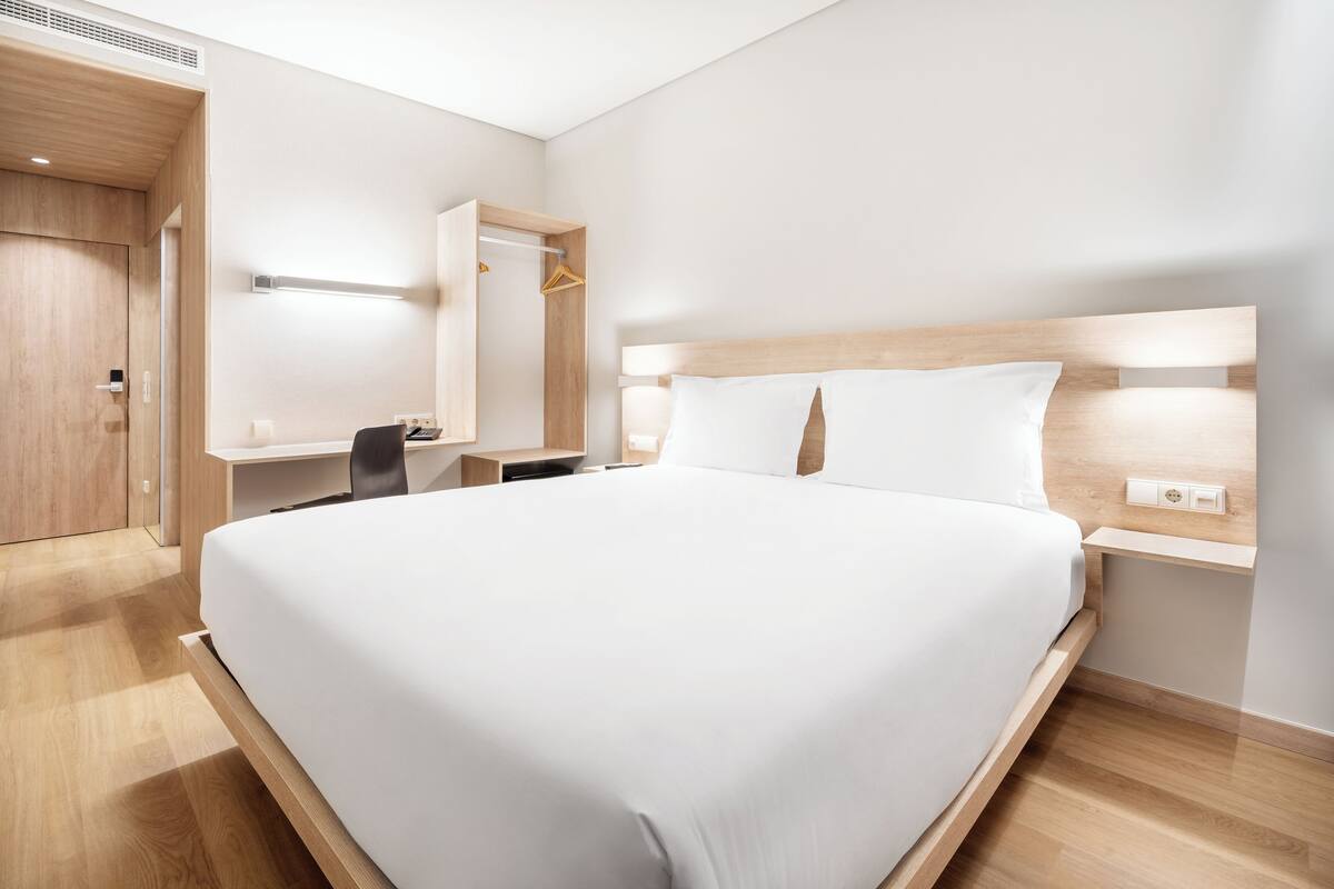 Une chambre d’hôtel moderne avec un grand lit, une tête de lit en bois, une table de chevet, un bureau avec une chaise et une armoire.