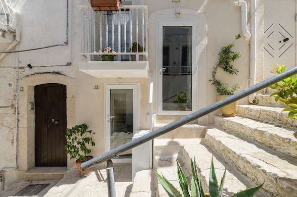 A narrow courtyard with a wooden door, a white door, and a balcony with potted plants.