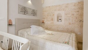 A bedroom with a bed, a headboard, a nightstand, a wall-mounted shelf, a decorative painting, and a stone wall with a niche.