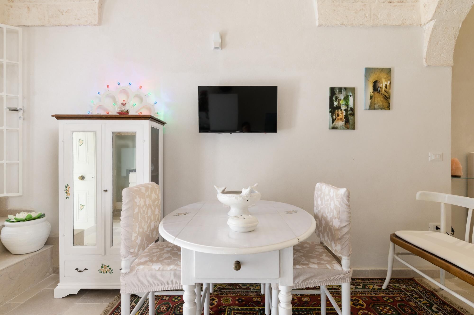 A dining area with a white round table, two chairs, a cabinet with floral designs, a television, and two framed pictures on the wall.