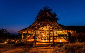 A thatched-roof structure with a wooden deck and glass walls, surrounded by dry grass and trees. - MISENI RETREAT (Saadani)