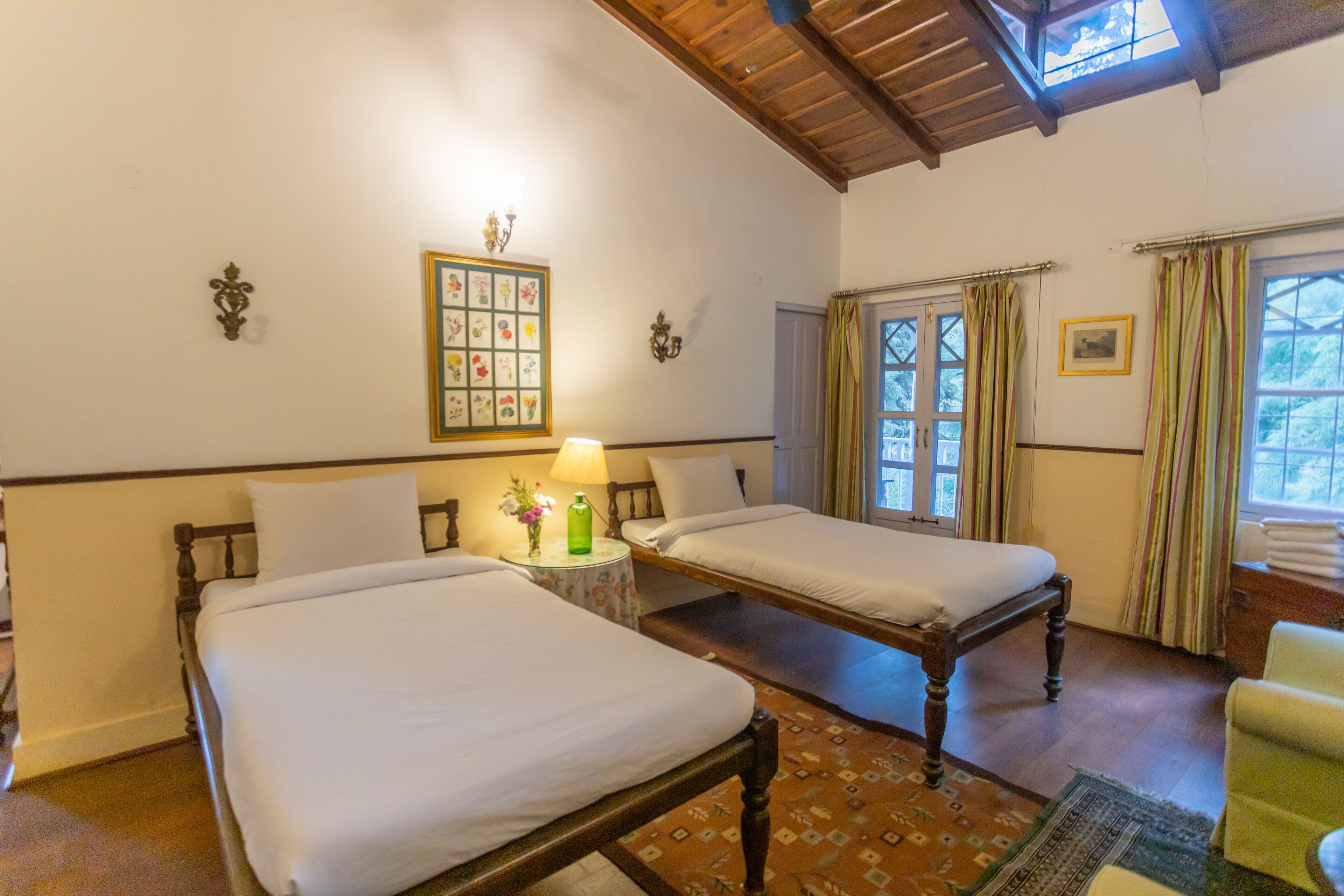 A hotel room with two beds, a wooden ceiling, a floral wall decoration, and a window with curtains.
