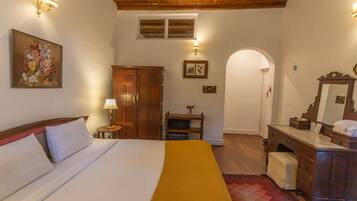 A hotel room with a bed, wooden wardrobe, a vanity with a mirror, and a painting on the wall.