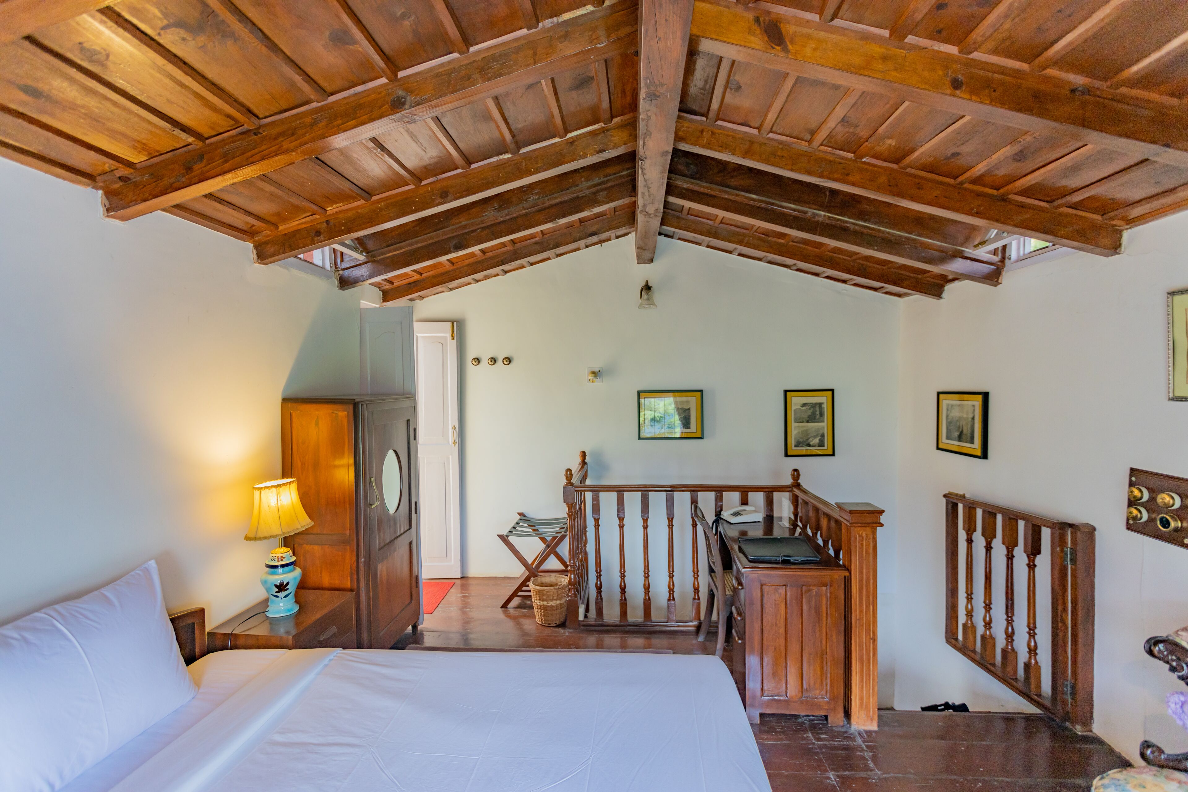 Une chambre avec un plafond en bois, un lit avec une literie blanche, une table de chevet avec une lampe, un bureau en bois et un escalier menant à un étage supérieur.