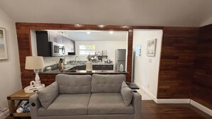 A living room with a grey sofa, wooden flooring, and a kitchen visible through an open doorway.