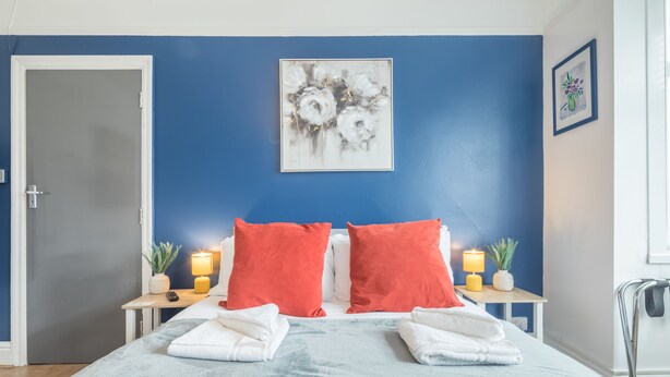 A bedroom with a bed, two red pillows, a blue wall, a framed artwork, and a bedside table with a lamp and a plant. - The Modern Cambridge Studio 4 (Cambridge)