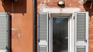 A tiled patio with a metal grate, a door with shutters, and air conditioning units mounted on a brick wall. - Bright Studio with Balcony by Wonderful Italy (Bologna)