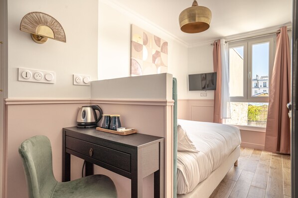 A bedroom with a bed, a desk with a kettle, a chair, a wall-mounted light, and a window with curtains.