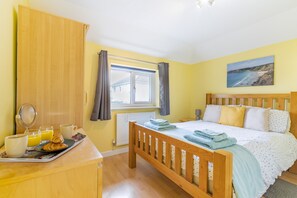 A bedroom with a wooden bed, a bedside table with a tray of croissants and orange juice, a window with curtains, and a painting of a beach on the wall. - Cornish Holiday - Meadow Rise (St Columb Major)