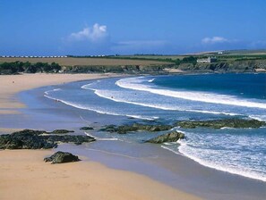 A beach with a curved shoreline, rocky outcrops, and a distant building. - Cornish Holiday - Meadow Rise (St Columb Major)