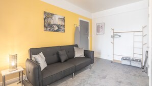 A modern living room with a black sofa, a small table with a lamp, a painting on the wall, and a drying rack with clothes. - The Modern Cambridge Studio 5 (Cambridge)