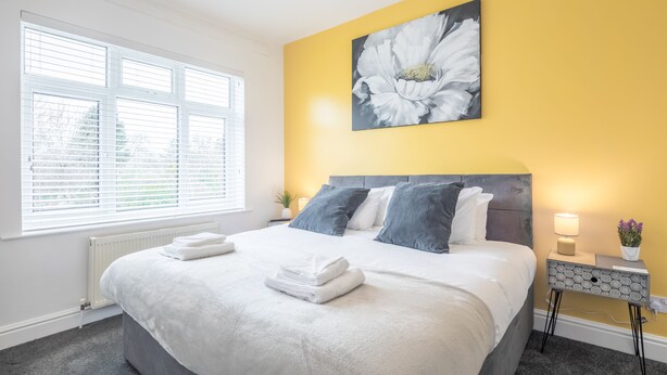 A neatly made bed with white linens, a large window with blinds, a wall-mounted flower painting, and a bedside table with a lamp and flowers. - The Modern Cambridge Studio 5 (Cambridge)