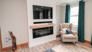 Un salón con un televisor de pantalla plana montado en una pared blanca, una chimenea debajo, un sillón capitonado con cojines y una mesa de centro de madera.