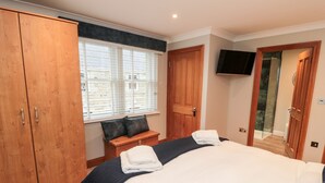 Un dormitorio con una cama grande, un armario de madera, un banco con cojines, un televisor montado en la pared y una puerta que da a un baño.
