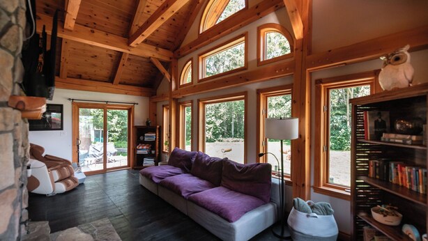 A cozy room with a stone wall, a purple sofa, a bookshelf, and large windows. - Timber Sky Estate - Family & Pet-Friendly Log Cabin in Elora (West Montrose)