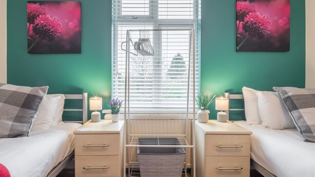 A bedroom with two beds, a window with blinds, a bedside table with a lamp, and a wall-mounted shelf. - The Modern Cambridge Studio 6 (Cambridge)