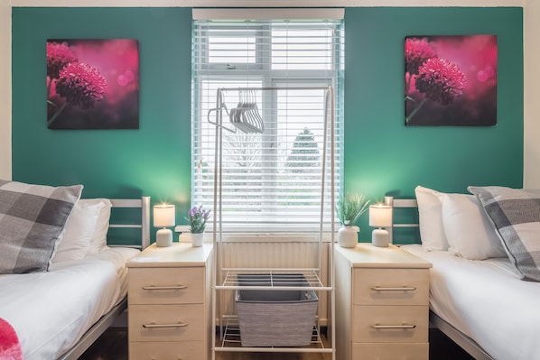 Two single beds with white bedding and grey checkered pillows, each flanked by a bedside table with a lamp and a small potted plant.