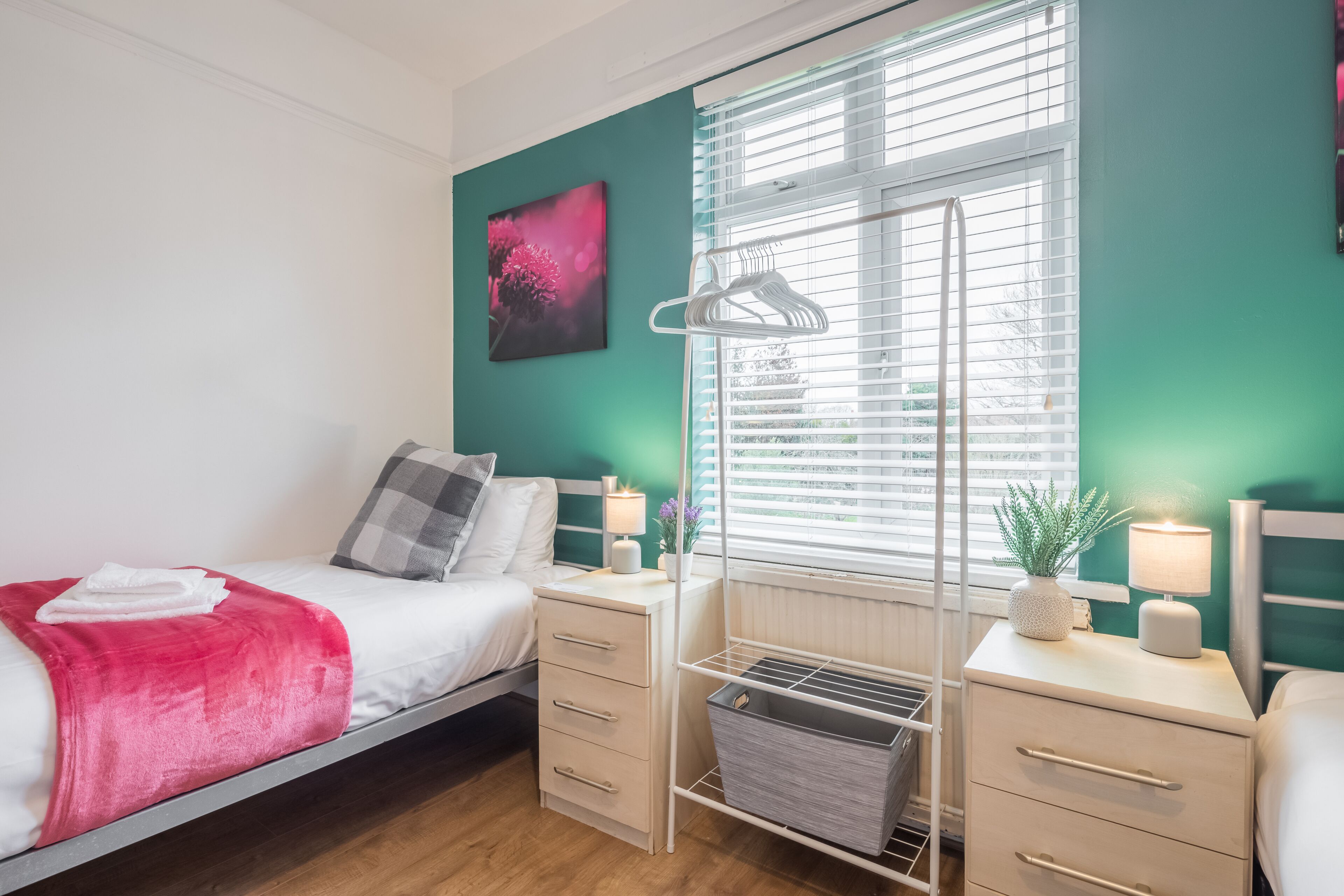 Un dormitorio con cama, mesitas de noche, radiador, ventana con persianas, una obra de arte en la pared y un perchero.