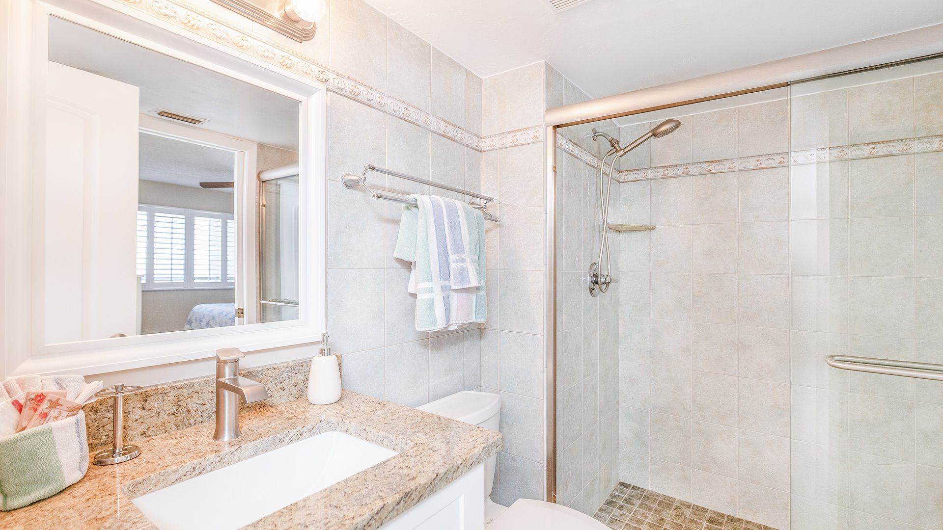 Un baño con un espejo grande, ducha con puerta de vidrio, encimera de granito y retrete.