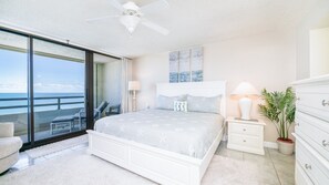 Quarto com cama, mesas de cabeceira, cômoda, ventilador de teto, vista para o oceano e uma cadeira perto da janela.