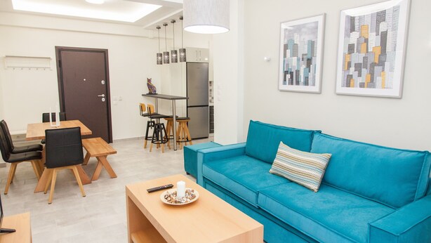 A modern living room with a teal sofa, wooden coffee table, and a dining area with a wooden table and chairs. - Supreme Athens Home by Cloudkeys (Zografos)