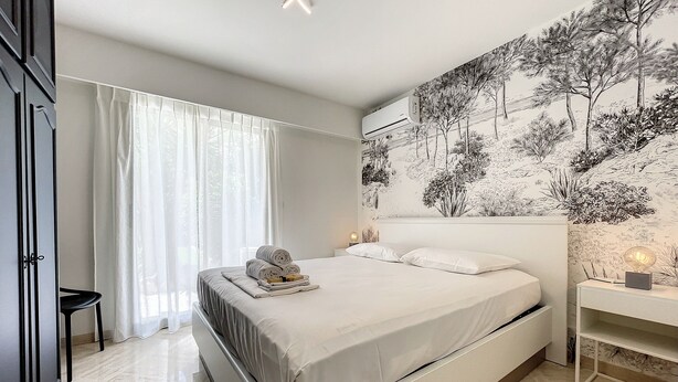 A bedroom with a bed, a nightstand, a wall-mounted air conditioner, and a window with sheer curtains. - (003)  Helena Garden City Trip (Antibes)