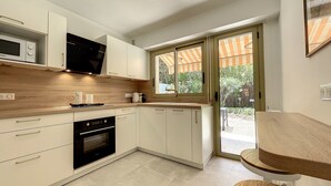 A modern kitchen with white cabinets, a built-in oven, and a microwave. There is a window with an orange-striped awning, and a wooden dining table with chairs. - (003)  Helena Garden City Trip (Antibes)