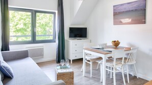 A room with a sofa, a dining table with chairs, a TV on a stand, and a painting on the wall. - Cozy stay for 2, terrace, 400m from beach (Fouesnant)