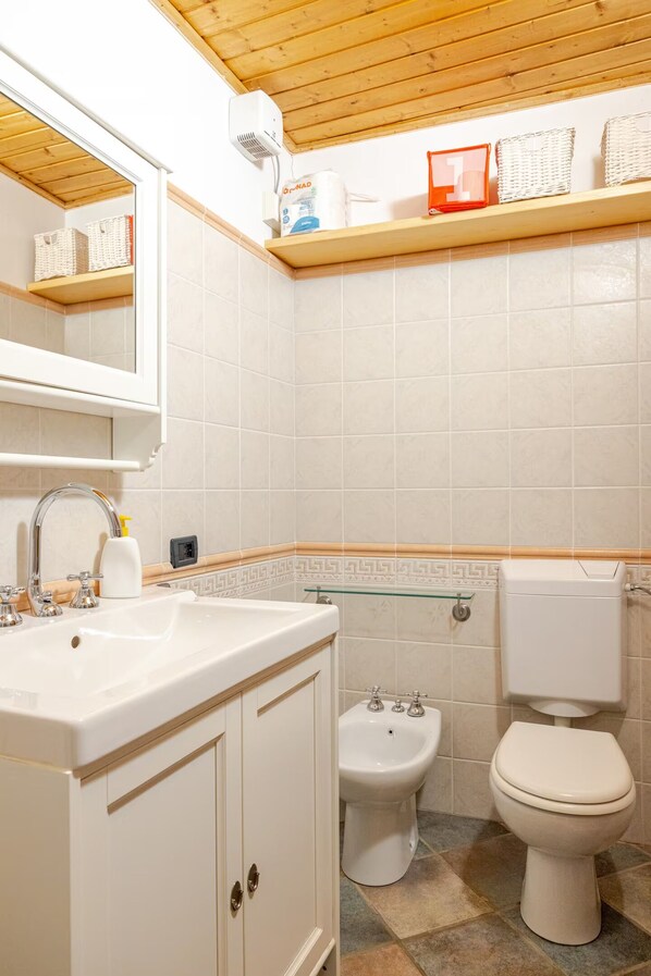 A bathroom with a white sink, a bidet, and a toilet. There is a mirror above the sink and a shelf with baskets and toiletries.