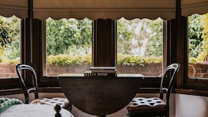 A room with a wooden table, two chairs, and a view of greenery through large windows. - Green Gables King Suite (Castlemaine)