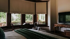 A bedroom with a bed, a television, a desk, and large windows with blinds. - Green Gables King Suite (Castlemaine)