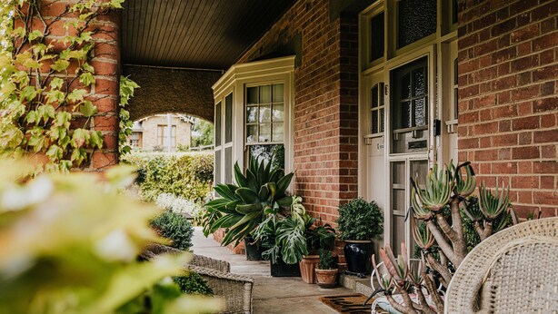 A covered porch with a brick wall, potted plants, and wicker furniture. - Green Gables King Suite (Castlemaine)