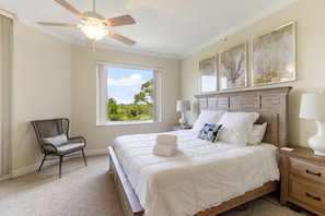 Quarto com cama, poltrona, mesa de cabeceira, ventilador de teto e janela com vista para vegetação.
